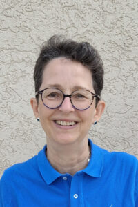 Man sieht Oberkörüer und Gesicht einer Frau mit Brille und dunklen kurzen Haaren.