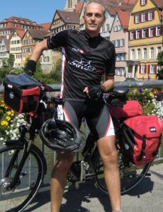 Mann in Radlerkluft lehnt vor seinem Rad, im Hintergrund Häuser