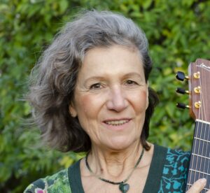 Porträtfoto der Kursleiterin: graue gewellte Haare, Gitarre in der Hand.