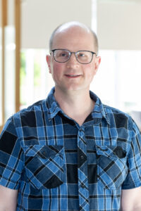 Mann mit kariertem blauen Hemd, der freundlich lächelt und eine Brille auf hat.