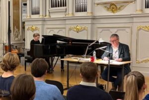 Eine Frau sitzt an einem Flügel und ein Mann an einem Tisch und liest vor.