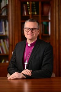 Bischof mit Kreuz um den Hals und Brille vor Bücherregal